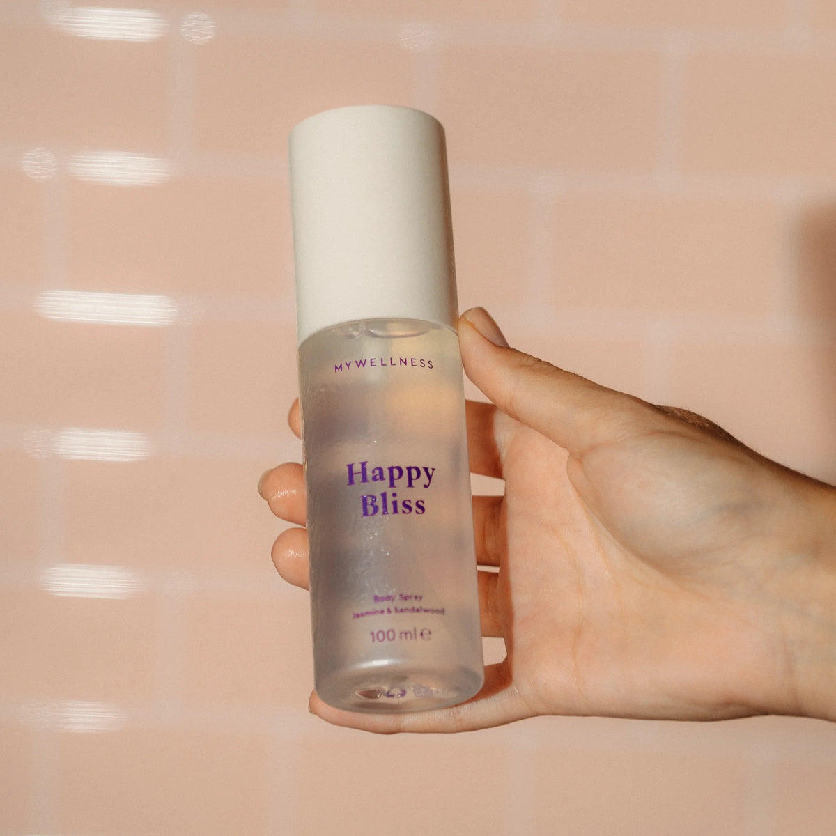 Hand hält Happy Bliss Body Spray mit Jasmin und Sandelholz vor rosa gefliester Wand