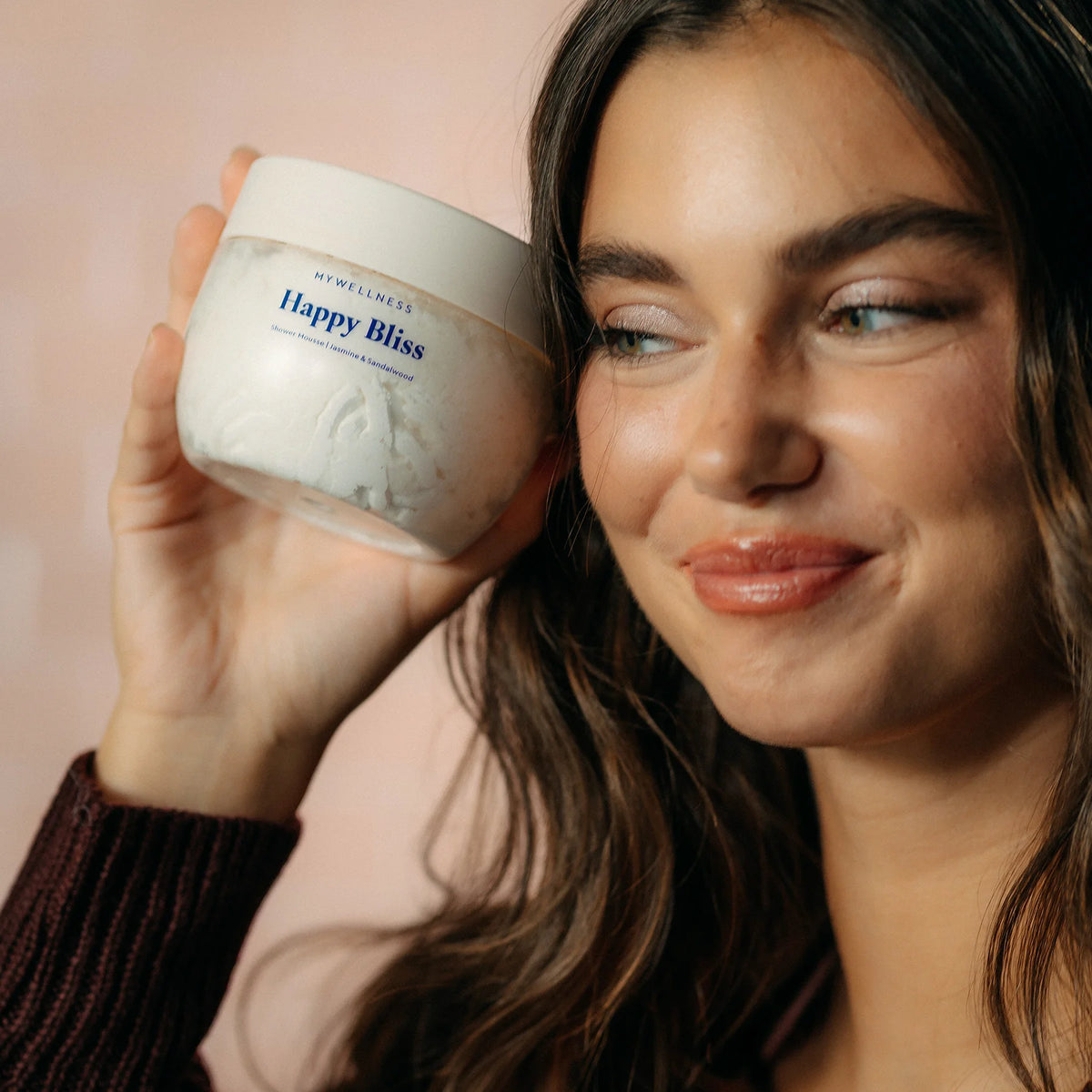 Lächelnde Frau im dunklen Strickpullover hält Happy Bliss Shower Mousse neben ihr Gesicht