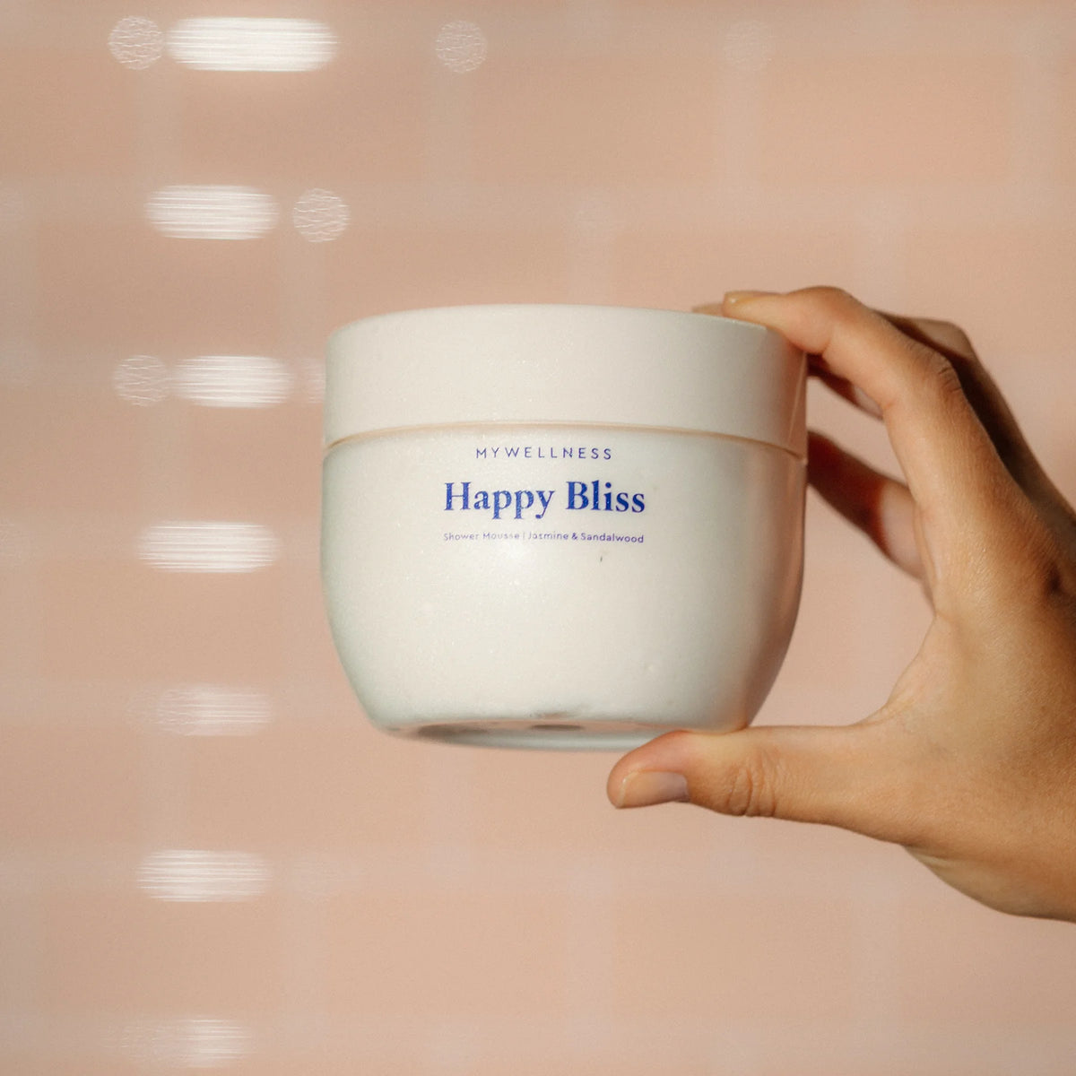 Hand hält Happy Bliss Shower Mousse mit Jasmin und Sandelholz vor rosa gefliester Wand