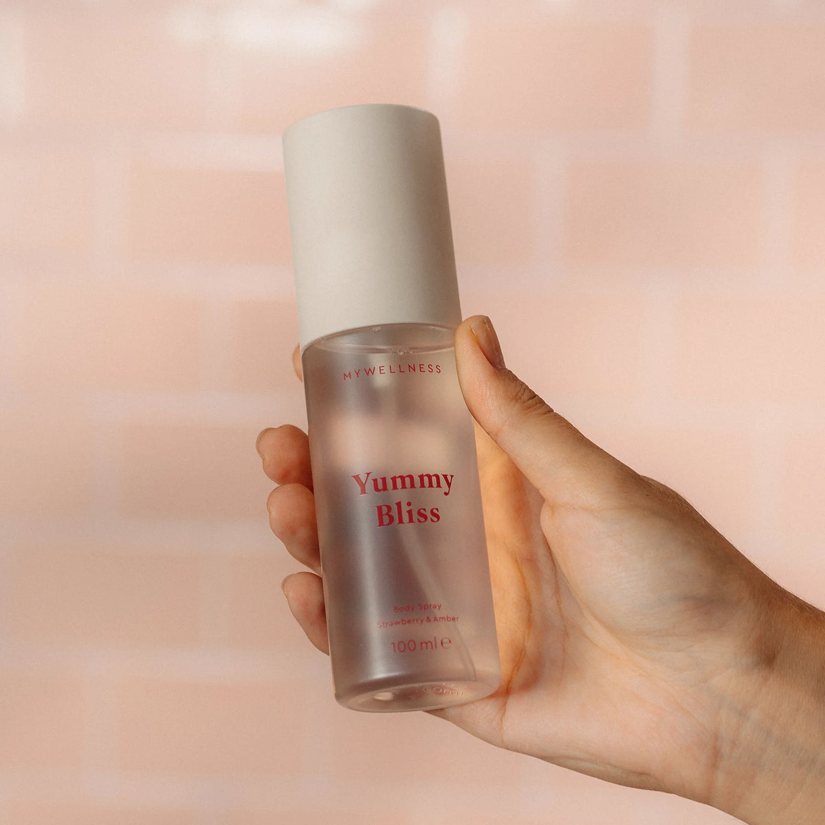 Hand hält Yummy Bliss Body Spray mit Erdbeere und Amber vor rosa gefliester Wand