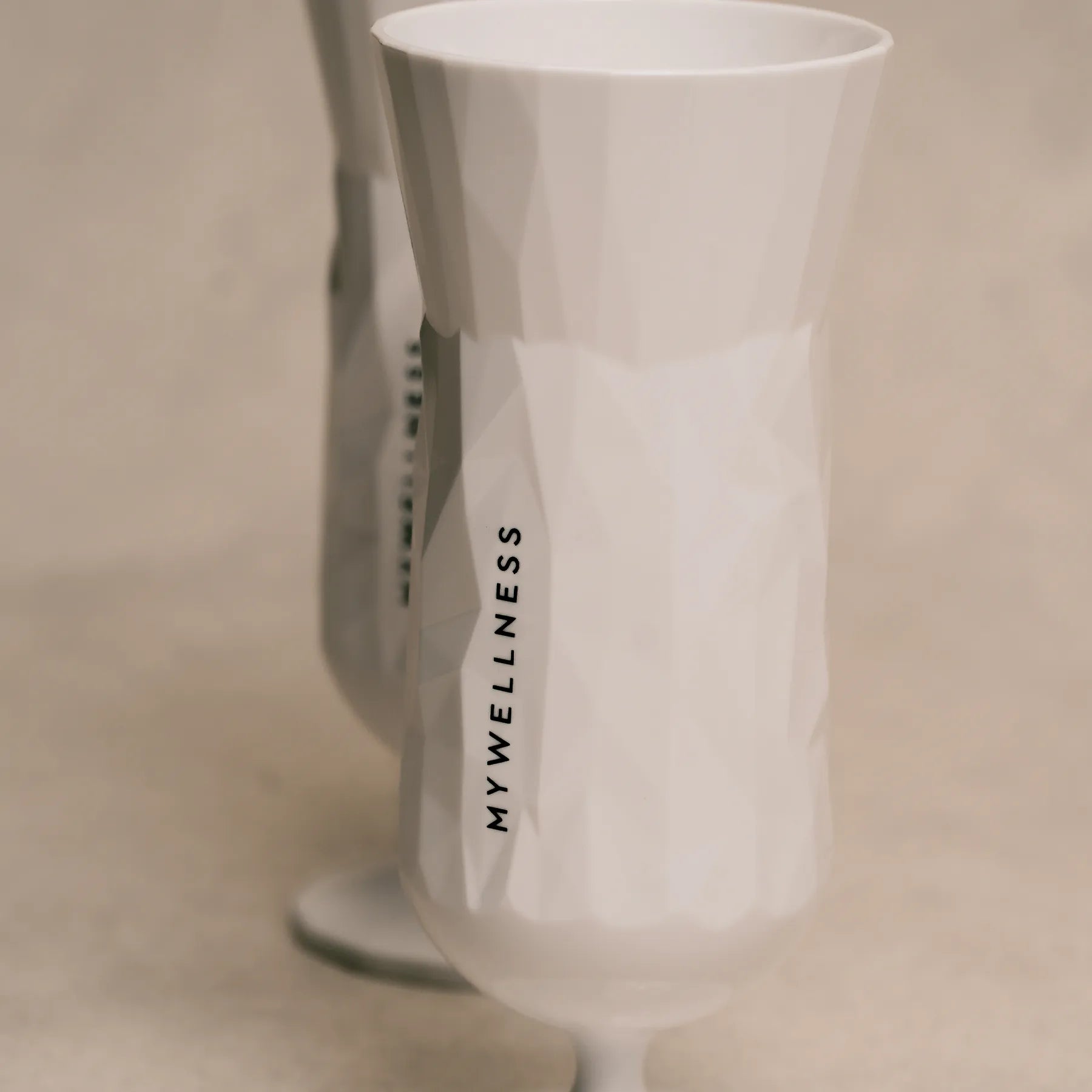 Weißer MYWELLNESS-Becher mit strukturierter Oberfläche und schwarzem Logo auf beigem Hintergrund.
