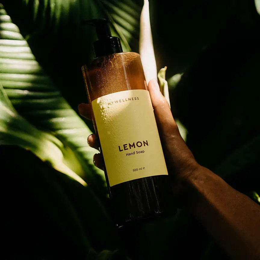 MyWellness Hand Soap Lemon in brauner Flasche – natürliche Handseife mit Zitronenduft, fotografiert im Sonnenlicht zwischen grünen Blättern in tropischer Umgebung