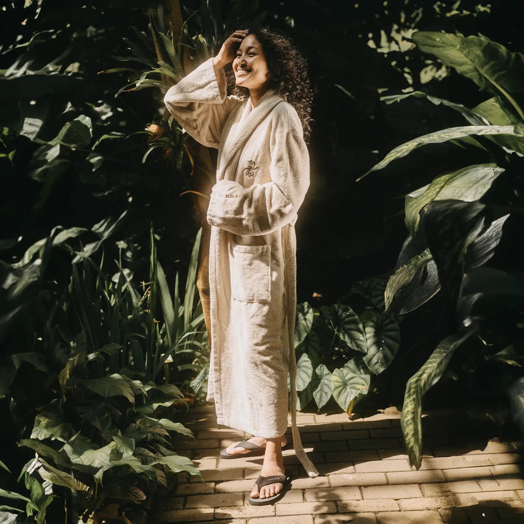 Frau in beigem MyWellness Bademantel in tropischer Umgebung – luxuriöse Spa-Ästhetik mit natürlichem Licht