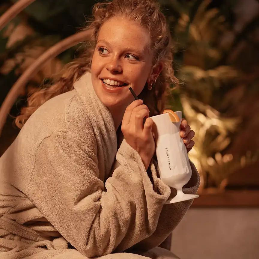 Frau in beigem Bademantel hält einen weißen MYWELLNESS-Becher und lächelt entspannt in warmer Wellness-Atmosphäre.