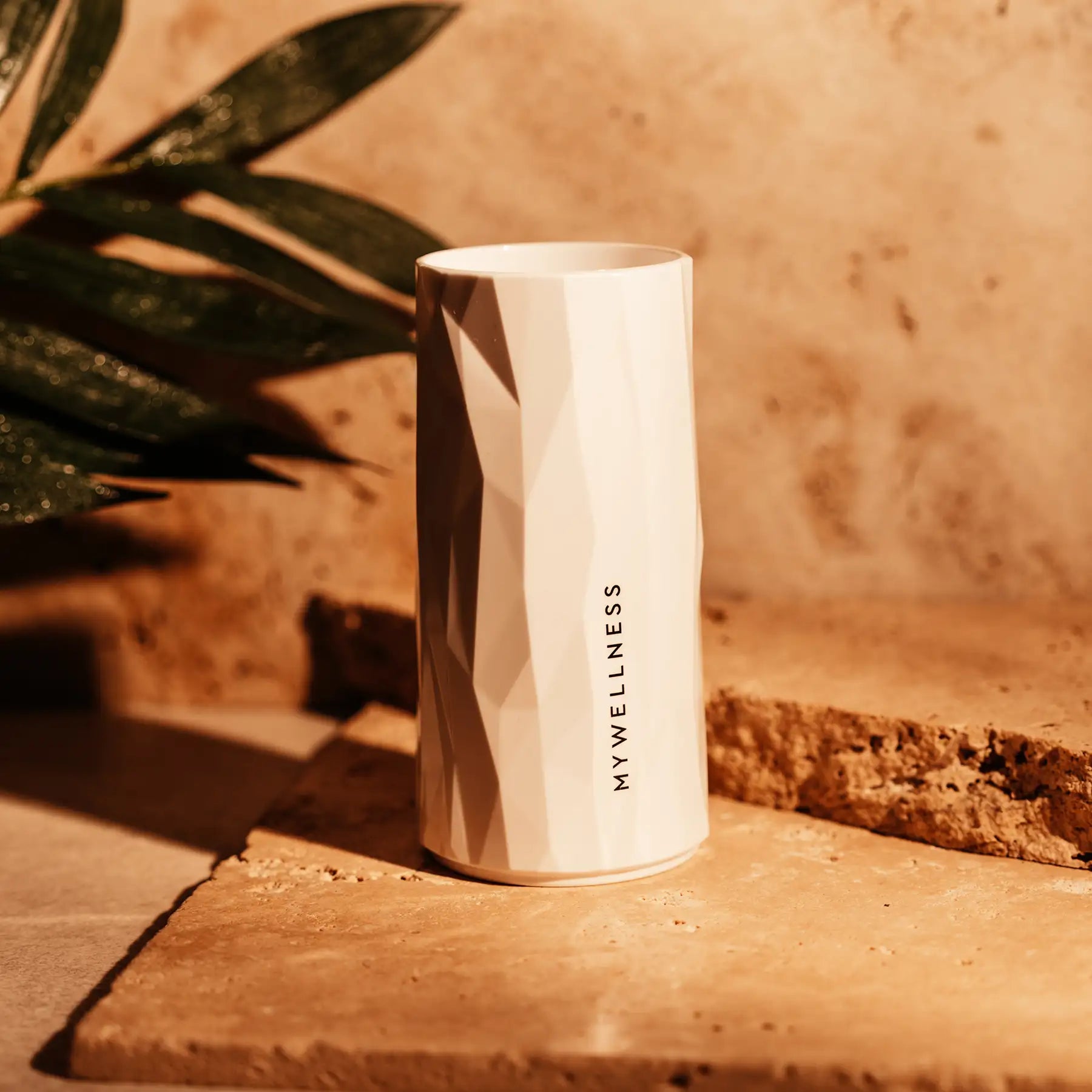 Weißes MYWELLNESS-Longdrinkglas mit strukturierter Oberfläche auf Natursteinuntergrund im Sonnenlicht.