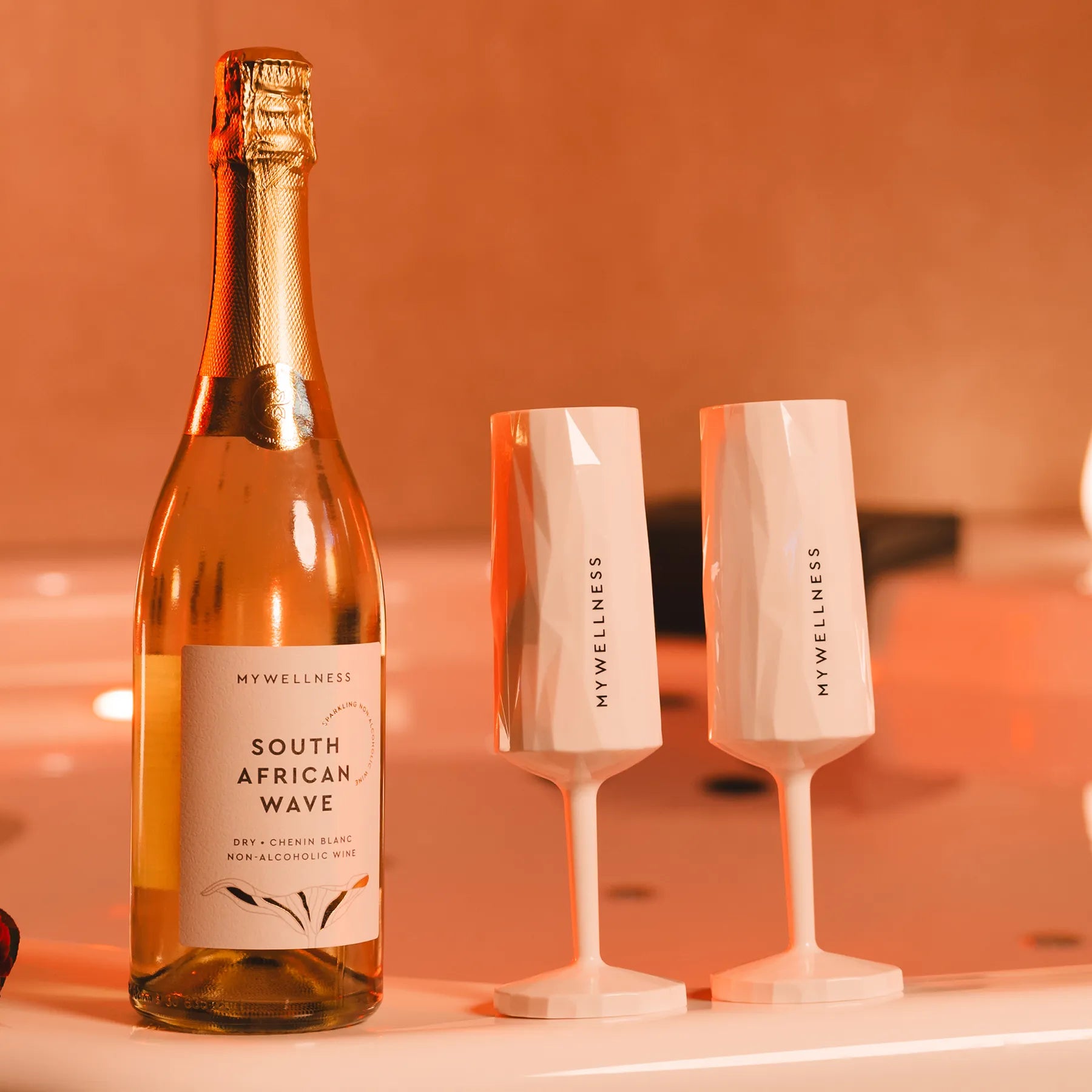 Zwei weiße MYWELLNESS-Sektgläser neben einer Flasche Sekt auf Natursteinuntergrund, beleuchtet von warmem Licht.