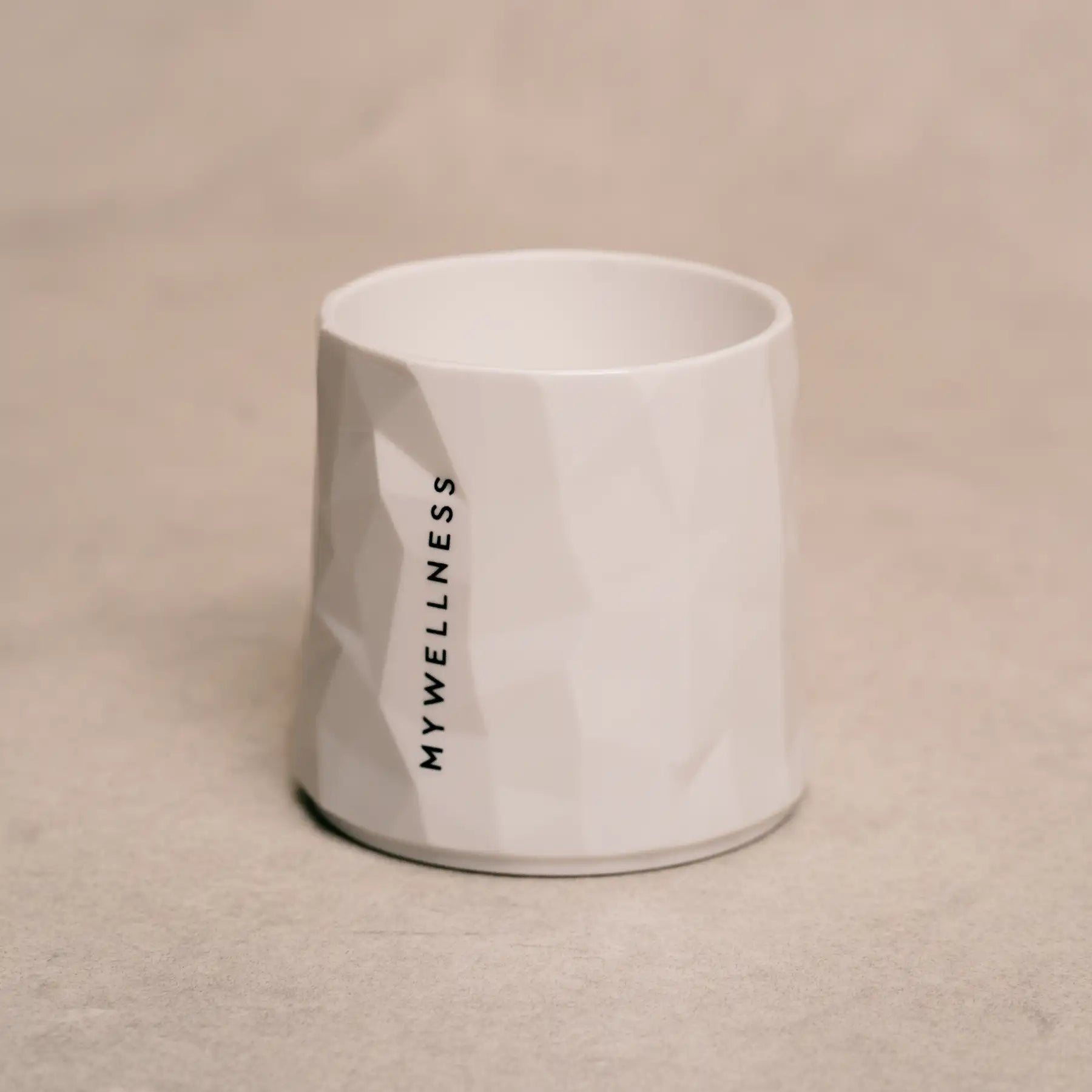 Weißer MYWELLNESS-Becher mit geometrischer Struktur und vertikalem Schriftzug, Studioaufnahme auf beigem Hintergrund.