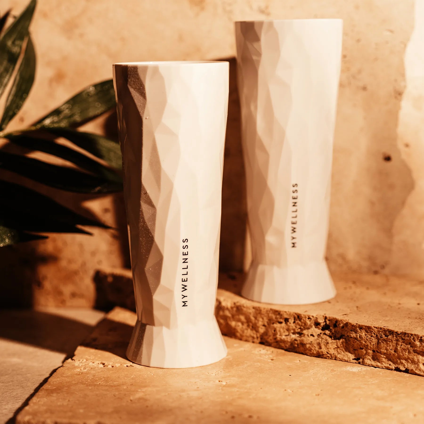 Weißes MyWellness-Weizenglas mit strukturierter Oberfläche, beleuchtet von warmem Sonnenlicht auf Steinuntergrund.